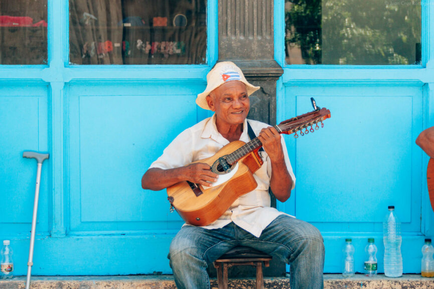 A smiling senior man sits on a stool playing a classic guitar in front of a bright light-blue wooden wall. He is wearing a white shirt, jeans, and a hat with a small flag detail. A walking cane and water bottles are visible beside him on the sidewalk.