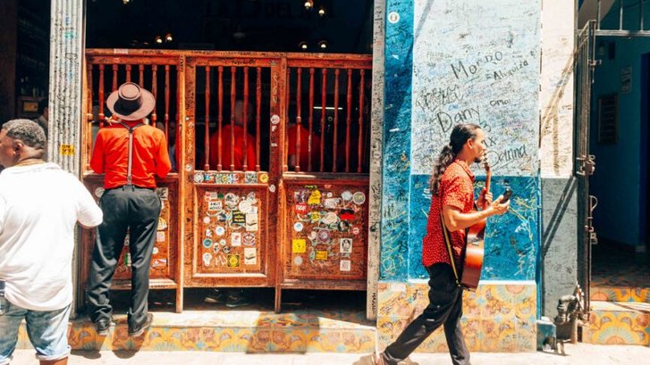 A man carrying an acoustic guitar walks past the entrance of a historic bar with wooden swinging doors covered in stickers. To the right, a bright blue wall is covered in handwritten graffiti and names. Another man in a red shirt and hat stands by the entrance