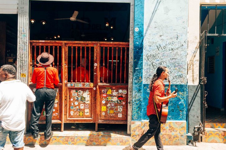 A man carrying an acoustic guitar walks past the entrance of a historic bar with wooden swinging doors covered in stickers. To the right, a bright blue wall is covered in handwritten graffiti and names. Another man in a red shirt and hat stands by the entrance
