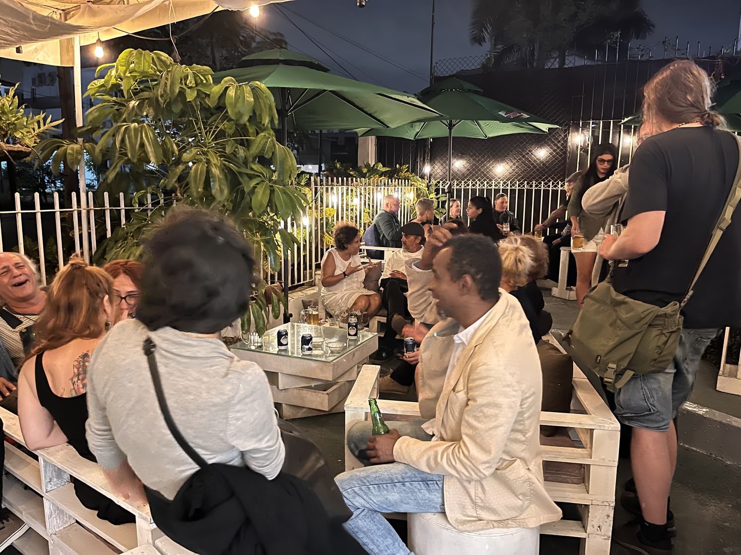 A lively outdoor terrace at Fellini in Miramar during the night, with people sitting on white wooden pallet furniture, drinking and talking under green umbrellas and string lights.